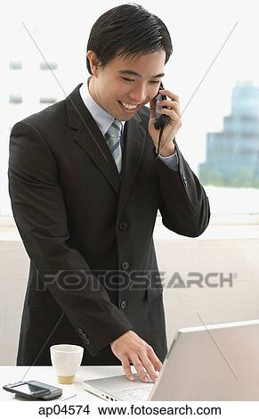 Picture - Businessman standing in office, using mobile phone and laptop, smiling. Fotosearch