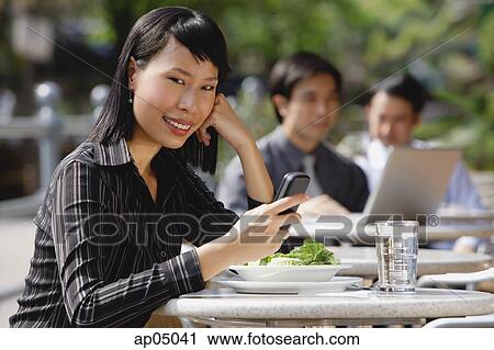 Stock Image - Businesswoman using mobile phone at outdoor cafe, text messaging, smiling at camera. Fotosearch