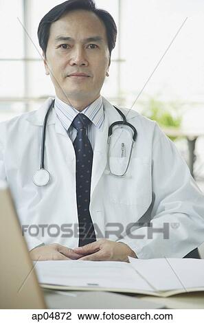 Doctor sitting at desk, looking at camera, serious expression View Large Photo Image Stock Image - Doctor sitting at desk, looking at camera, serious expression. Fotosearch