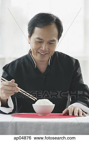 Stock Photograph - Man looking at bowl of rice on table, holding chopsticks. Fotosearch