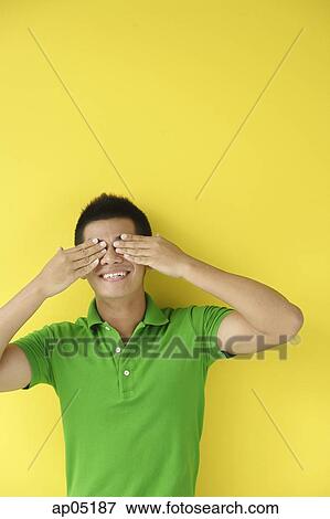 Stock Photo - Man standing against yellow wall, covering eyes. Fotosearch