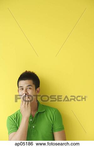 Stock Photograph - Man standing against yellow wall, covering mouth. Fotosearch