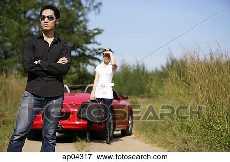 Man standing with arms crossed, woman and red sports car in the background View Large Photo Image Stock Photo - Man standing with arms crossed, woman and red sports car in the background. Fotosearch