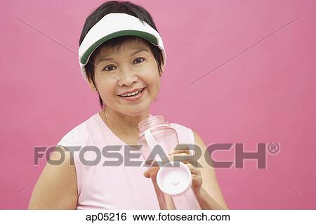 Stock Photograph - Mature woman wearing sun visor and water bottle, smiling. Fotosearch