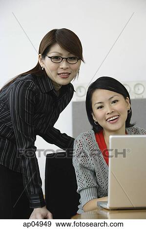 Stock Photo - Two businesswomen, smiling at camera, laptop open in front of them. Fotosearch