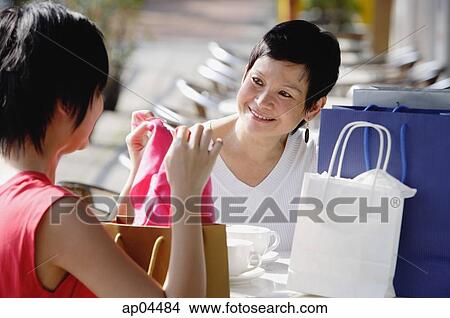 Two women at cafe, looking at items from their shopping bags View Large Photo Image Picture - Two women at cafe, looking at items from their shopping bags. Fotosearch