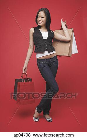 Stock Photo - Woman carrying shopping bags, standing against red wall. Fotosearch
