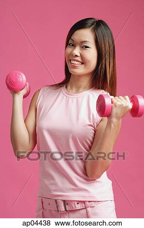 Stock Photo - Woman dressed in pink, standing against pink wall, holding dumbbells. Fotosearch