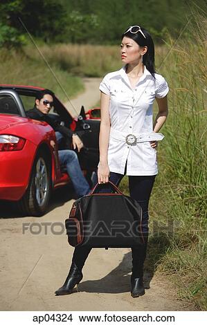 Picture - Woman in foreground carrying bag, man sitting in red sports car in the background. Fotosearch