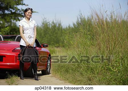 Stock Photo - Woman standing next to red sports car, carrying bag. Fotosearch