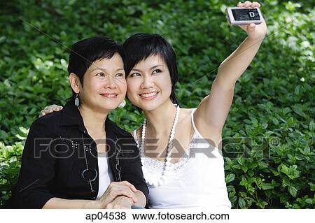 Stock Photo - Women side by side, smiling, taking a picture with mobile phone. Fotosearch
