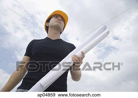 Stock Photo - Young man wearing hardhat, carrying blueprints, low angle view. Fotosearch