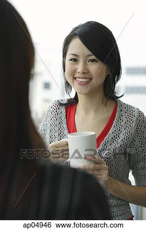 Stock Photograph - Young woman holding mug, smiling at person in front of her. Fotosearch