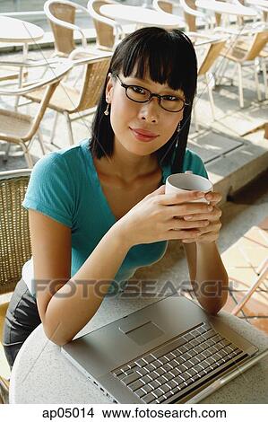 Young woman in cafe, holding cup, laptop on table next to her, looking at camera View Large Photo Image Picture - Young woman in cafe, holding cup, laptop on table next to her, looking at camera. Fotosearch