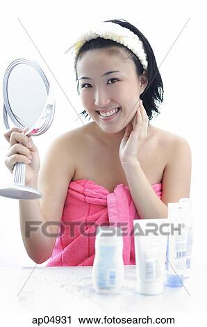 Stock Image - Young woman sitting at dressing table, hand on chin, holding mirror. Fotosearch