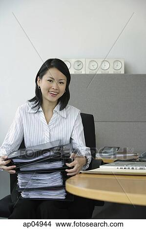 Picture - Young woman sitting at office desk, holding stack of binders. Fotosearch