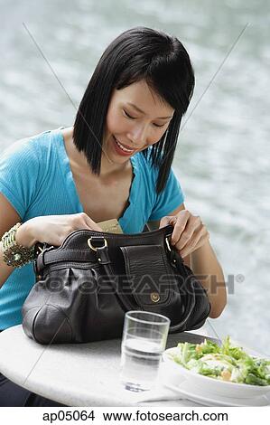 Picture - Young woman sitting at riverside cafe, looking through her handbag. Fotosearch