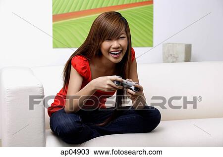 Stock Image - Young woman sitting on sofa, holding TV remote control. Fotosearch