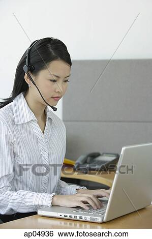 Stock Photograph - Young woman using laptop, wearing hands free device. Fotosearch