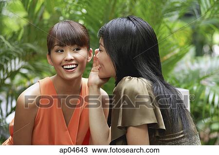 Stock Image - Young woman whispering in ear of another woman. Fotosearch