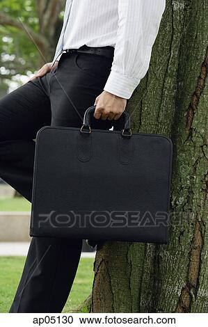 Stock Image - Businessman leaning on tree, holding briefcase, cropped image. Fotosearch