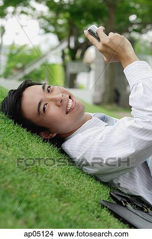 Businessman lying on grass in park, holding mobile phone, smiling at camera View Large Photo Image Picture - Businessman lying on grass in park, holding mobile phone, smiling at camera. Fotosearch