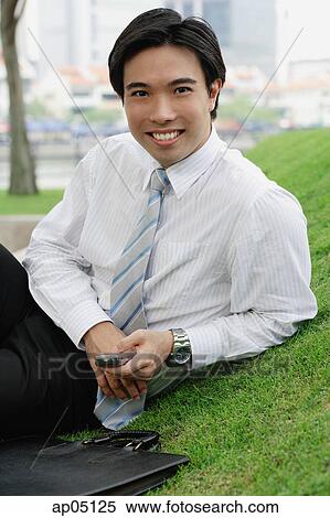 Businessman sitting on grass in park, holding mobile phone, smiling at camera View Large Photo Image Stock Photography - Businessman sitting on grass in park, holding mobile phone, smiling at camera. Fotosearch