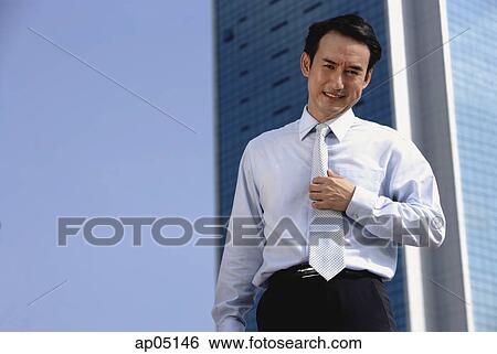 Stock Photograph - Businessman standing looking at camera, hand on tie. Fotosearch