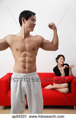 Couple at home, man in foreground showing off muscles, woman in background, sitting on sofa View Large Photo Image Stock Image - Couple at home, man in foreground showing off muscles, woman in background, sitting on sofa. Fotosearch