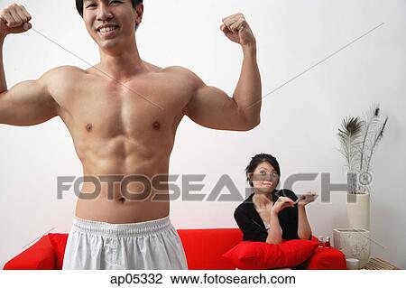 Couple at home, man in foreground flexing muscles, woman sitting on sofa View Large Photo Image Stock Image - Couple at home, man in foreground flexing muscles, woman sitting on sofa. Fotosearch