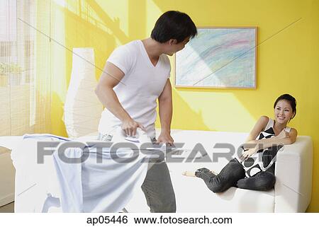 Couple at home, man ironing turning to look at woman sitting on sofa behind him View Large Photo Image Stock Photograph - Couple at home, man ironing turning to look at woman sitting on sofa behind him. Fotosearch
