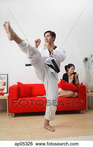 Stock Photograph - Couple at home, man practicing martial arts, woman sitting on sofa, filing her nails. Fotosearch