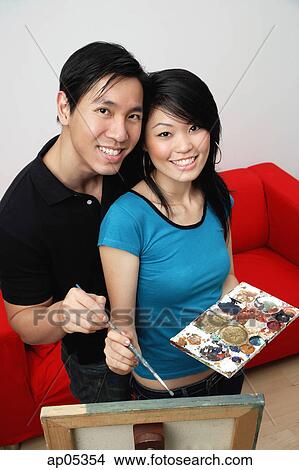 Picture - Couple at home, standing in front of easel with palette and paint brushes, smiling at camera. Fotosearch