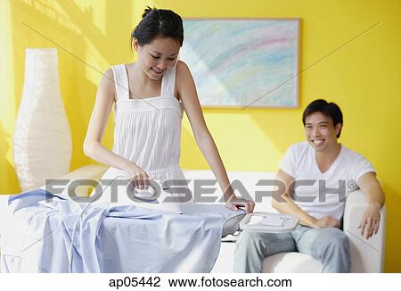 Stock Image - Couple at home, woman ironing, man sitting on sofa behind her. Fotosearch