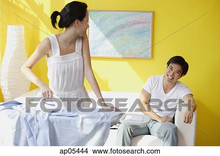 Couple at home, woman ironing turning to look at man sitting on sofa behind her View Large Photo Image Picture - Couple at home, woman ironing turning to look at man sitting on sofa behind her. Fotosearch