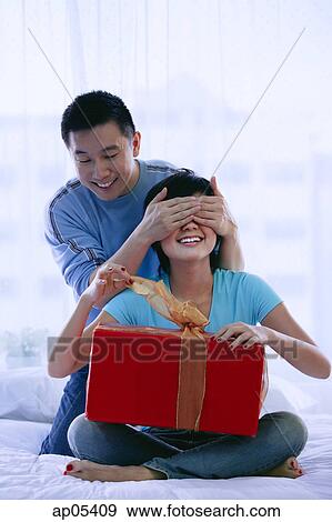 Stock Photo - Couple in bedroom, woman opening gift, man covering her eyes. Fotosearch