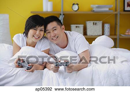 Couple lying on bed, holding video game controls, smiling at camera View Large Photo Image Stock Image - Couple lying on bed, holding video game controls, smiling at camera. Fotosearch