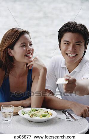 Stock Photo - Couple sitting at outdoor cafe, man holding credit card towards camera. Fotosearch