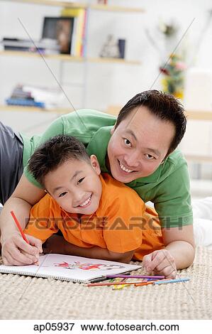 Stock Photo - Father and son at home, lying on floor with sketch pad, smiling at camera. Fotosearch