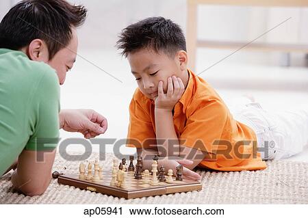 Stock Image - Father and son lying on floor, playing chess. Fotosearch