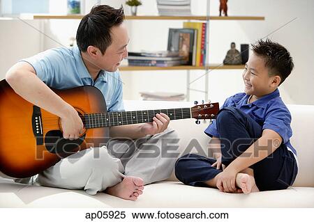 Stock Photography - Father and son sitting on sofa, father playing guitar. Fotosearch