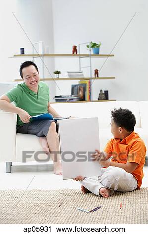 Stock Image - Father sitting on sofa, son on floor holding up drawing pad. Fotosearch