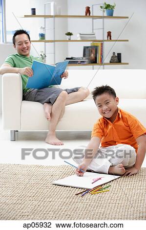 Stock Image - Father sitting on sofa, son on floor with drawing pad. Fotosearch