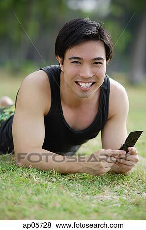 Man lying on grass, holding mobile phone, smiling at camera View Large Photo Image Stock Photo - Man lying on grass, holding mobile phone, smiling at camera. Fotosearch