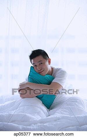 Stock Photo - Man sitting in bed, embracing pillow, smiling at camera. Fotosearch