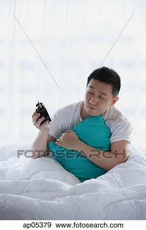 Man sitting in bed, holding alarm clock View Large Photo Image Stock Photo - Man sitting in bed, holding alarm clock. Fotosearch