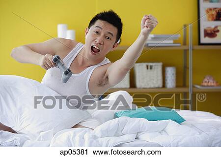 Stock Image - Man sitting in bed, holding video game remote control, arms outstretched. Fotosearch
