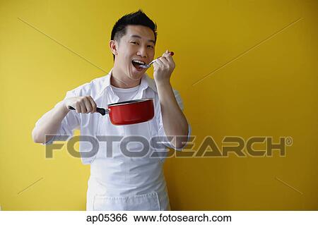 Stock Photograph - Man wearing apron and holding saucepan, eating from spoon. Fotosearch