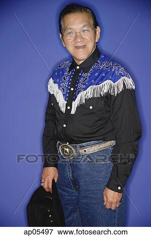 Stock Photo - Senior man dressed in cowboy attire, standing against blue background, portrait. Fotosearch