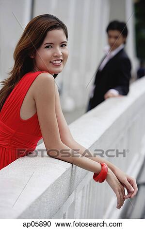 Stock Image - Woman leaning on bridge railing, smiling at camera, man in the background. Fotosearch